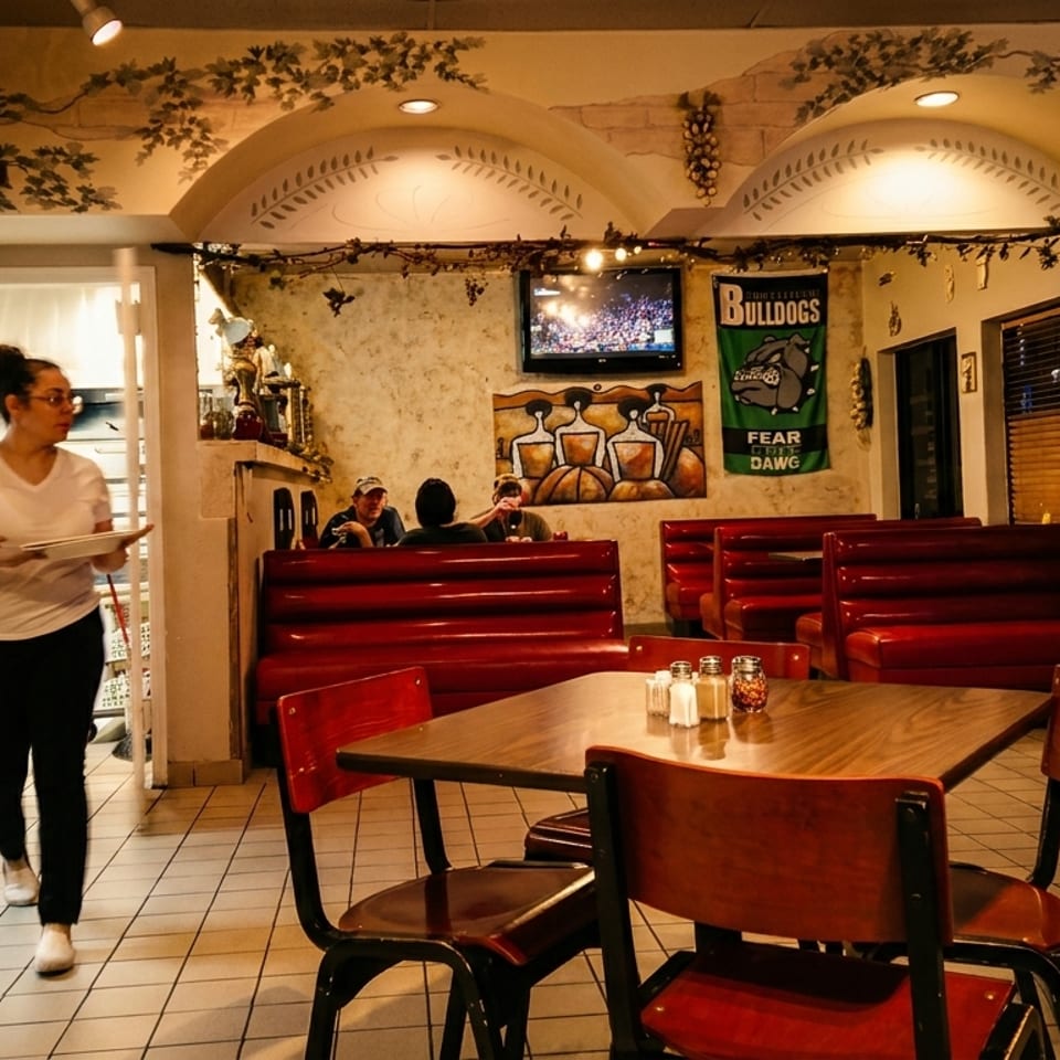 Warmly lit restaurant dining room with red booths, a server carrying plates, wall art, television, and candy machine.